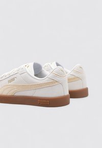 Zapatillas de ante blancas con una franja de cuero marrón y suela de goma marrón. Presenta un logo dorado en el lateral y el talón. Diseño de punta redonda.