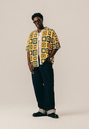 Hombre con gafas, camisa de manga corta con estampado amarillo y verde, camiseta blanca debajo, vaqueros oscuros con puños y zapatos negros, de pie con una mano en el bolsillo.