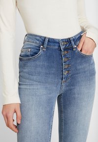 Pantalones vaqueros de mezclilla azul de talle alto con una bragueta de cinco botones, bolsillos frontales y un ligero desvanecimiento, con costuras de cobre y una textura suave.