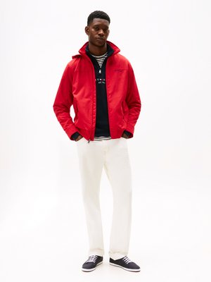 Casaco leve vermelho com colarinho alto, usado sobre um hoodie azul-marinho e uma camisa riscada. Combinado com calças brancas e ténis pretos.