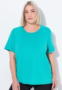 Blonďatá žena se rovnými vlasy, oblečená v tyrkysovém tričku s krátkým rukávem a černých kalhotách, stojící na bílém pozadí.