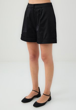 Zwarte satijnen shorts met een geplooide voorkant, omgeslagen zoom en een zachte textuur. Gecombineerd met zwarte flats met dunne enkelbandjes.