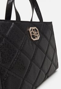 Zwarte gewatteerde draagtas van gestructureerd materiaal, met een gouden achthoekig logo-accent aan de voorkant en dubbele handvatten.