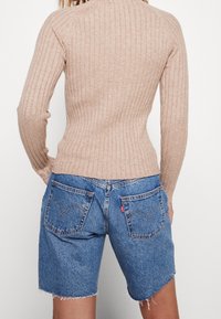 Beige gestreifter Pullover mit langen Ärmeln, kombiniert mit blauen Jeansshorts, die fransige Säume und Gesäßtaschen mit einem Stichmuster aufweisen.