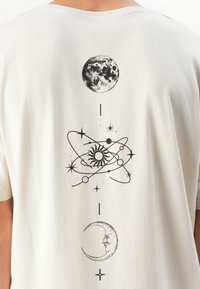 Black celestial design on a white shirt back showing a full moon, a sun with orbiting planets, and a crescent moon with a face.