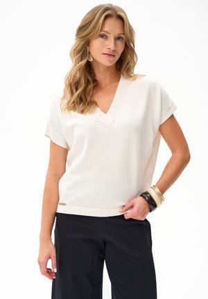 Blondynka w białej koszulce z krótkim rękawem i dekoltem w serek, czarnych spodniach oraz z nałożonymi na prawym nadgarstku bransoletkami, pozująca na białym tle.