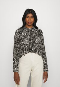 Blusa de mangas longas em estampado de leopardo preto e bege, com gola alta e detalhe franzido, combinada com calças de cor clara.