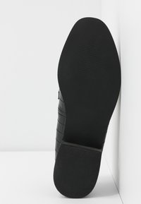 Scarpa nera in pelle con un motivo in croco testurizzato, suola piatta in gomma e punta liscia e arrotondata. La vista posteriore mette in evidenza il design della superficie anti-scivolo della suola.