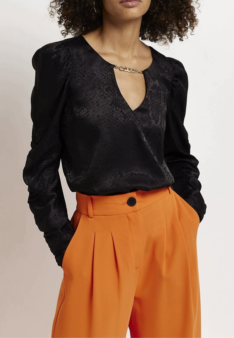 Camicia nera a maniche lunghe con un motivo testurizzato e scollo a V, accentuata da una catena dorata. Abbinata a pantaloni arancioni a vita alta e plissettati.