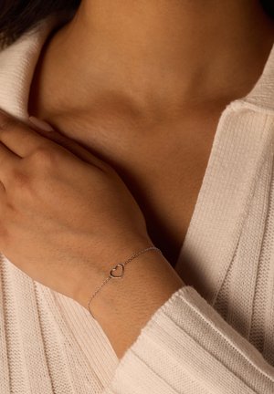 Zilveren armband met een hartvormige bedel, met een delicate ketting, tegen een gestructureerde trui in een zachte, neutrale kleur.