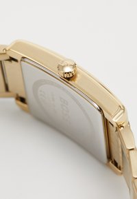 Montre-bracelet en ton or avec un boîtier rectangulaire, finition brossée et couronne texturée avec le logo "BOSS" gravé. Fond blanc avec détail de logo.