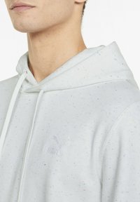 Joven con una sudadera ligera gris claro con manchas multicolores y un sutil logo de Puma en el pecho, mostrando el cuello y la parte inferior de la cara.