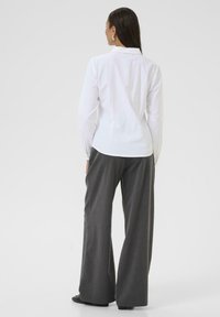 Chemise blanche à manches longues avec un col classique et un empiècement dans le dos, associée à un pantalon large gris et des chaussures noires. Tissu lisse et taillé.