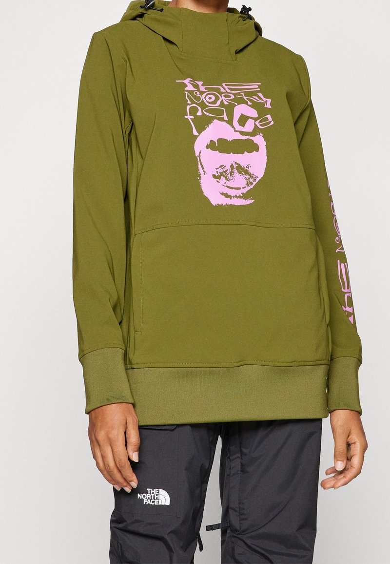 Personne portant un sweat à capuche vert olive avec un logo déformé rose « The North Face » et un pantalon noir avec un logo blanc « The North Face » sur la cuisse gauche.