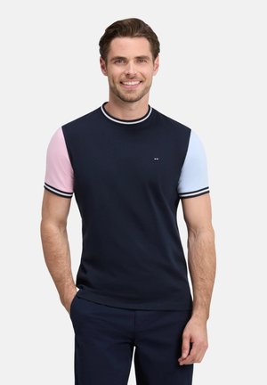 Homme souriant portant un t-shirt bleu marine avec une manche gauche rose et une manche droite bleu clair, mains dans les poches d'un pantalon bleu marine, fond blanc.