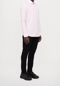 Camisa de manga larga a rayas en rosa claro con cuello, combinada con pantalones negros y zapatillas negras, mostrando una silueta ajustada.