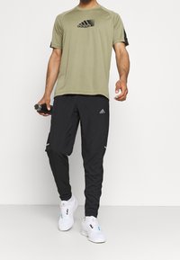 Olivgrünes Sportshirt mit kurzen Ärmeln und schwarzem Logo, kombiniert mit schwarzen Hosen mit reflektierenden Akzenten. Weiße Sportschuhe.