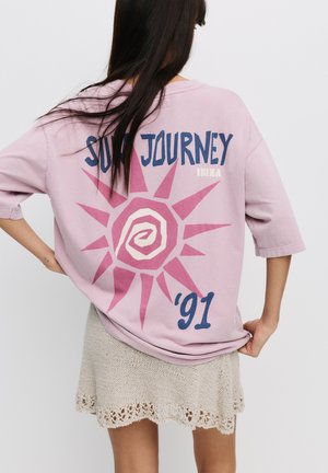 Roza oversize majica s grafiko sonca in natisnjenim besedilom 'SUN JOURNEY 91' v modri barvi, v kombinaciji z pleteno bež krilom z valovitimi robovi.