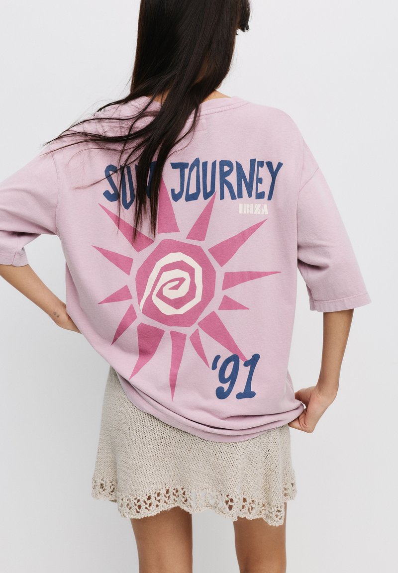 Lyserød oversized t-shirt med et solgrafik og teksten 'SUN JOURNEY 91' trykt i blå, samlet med en strikket beige nederdel med bølgede kanter.
