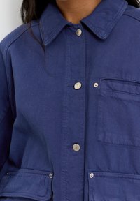 Navy blue jacket made of textured fabric, featuring a collared neckline, silver buttons, and a chest pocket with a flap.