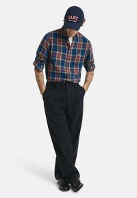 Geruit katoenen overhemd in blauw, groen en rood, met opgerolde mouwen. Donkere broek en glanzende zwarte schoenen. Pet met 'GANT' logo in het rood.