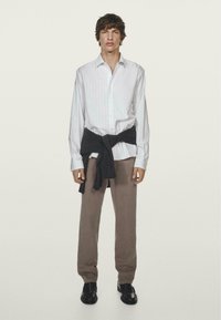 Weiß gestreiftes Hemd mit Knöpfen, graue Hosen und schwarze Schuhe. Dunkler Pullover um die Taille gebunden. Modell steht vor einem hellen Hintergrund.