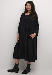 Black, knee-length dress with diamond pattern, long sleeves, and side pockets. Paired with black ankle boots, showcasing a relaxed fit.