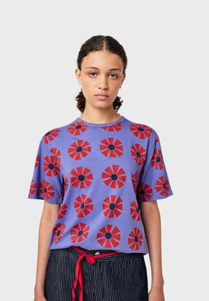 Lila Baumwoll-T-Shirt mit roten Blumenmustern und schwarzen Zentren, Rundhalsausschnitt und kurzen Ärmeln, getragen mit gestreiften Hosen und einem roten Gürtel.