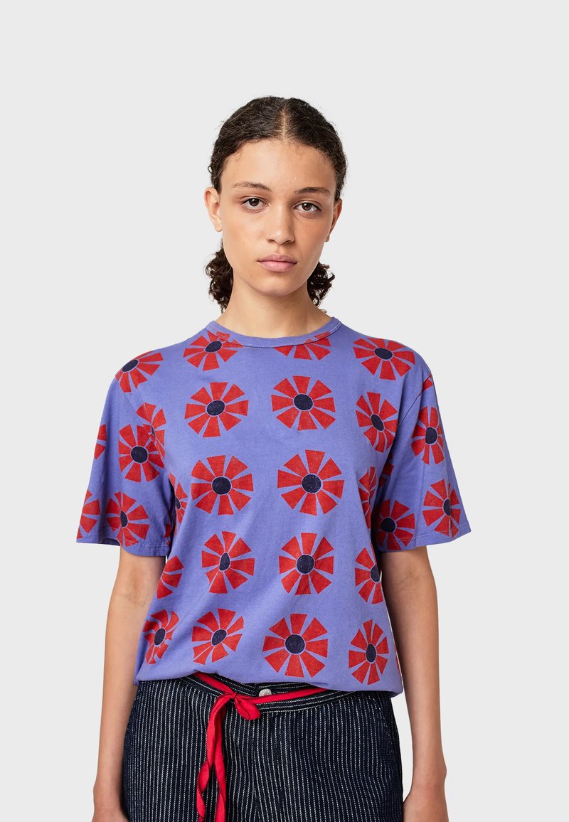 Camiseta de algodón morada con estampados de flores rojas y centros negros, cuello redondo y mangas cortas, combinada con pantalones de rayas y un cinturón rojo.