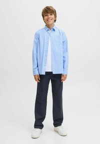 Lachende jongen die staat, met een lichtblauw gestreept overhemd over een wit T-shirt, donkere broek en witte sneakers op een effen witte achtergrond.