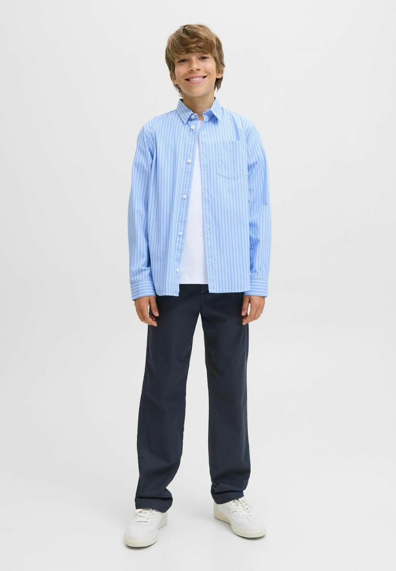 Lachende jongen die staat, met een lichtblauw gestreept overhemd over een wit T-shirt, donkere broek en witte sneakers op een effen witte achtergrond.
