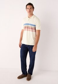 Polo color crema con righe orizzontali blu, marroni e rosa; maniche corte, design con colletto. Indossata con jeans blu scuro e scarpe marroni.