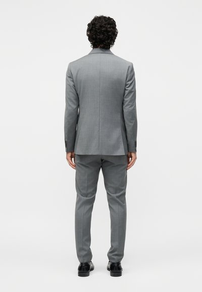 Άνδρας με σγουρά μαλλιά που στέκεται γυρισμένος πλάτη, φοράει εφαρμοστό γκρι κοστούμι με μαύρα επίσημα παπούτσια, πάνω σε απλό φόντο.