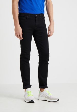Hombre vestido con jeans ajustados negros, zapatillas blancas con cordones verde neón y una camisa azul, de pie sobre un fondo blanco.