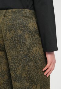 Παντελόνι σε ελαιόχρωμο πράσινο με μαύρο animal print σχέδιο, με πλαϊνές τσέπες και υφασμάτινη υφή, μερικώς ορατό πάνω από ένα μαύρο τοπ.