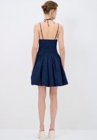 Robe bleu marine avec un corsage ajusté plissé et une jupe évasée plissée. Présente des bretelles spaghetti fines et un nœud dans le dos.