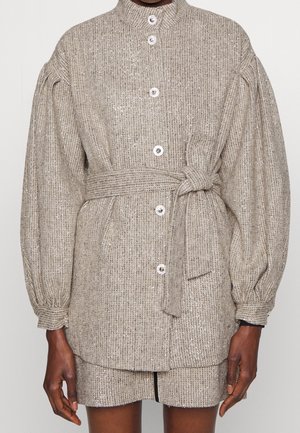 Chaqueta beige de manga larga con textura, botones frontales y cinturón a juego anudado en la cintura, llevada sobre una minifalda beige.