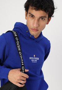 Haut en coton bleu avec une poche avant, capuche à cordon et logo "HILFIGER NEW YORK". La sangle noire porte l'inscription "HUGO HUGO".