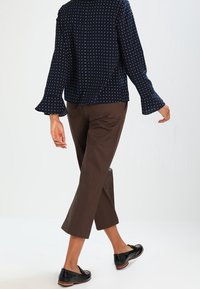 Persona che indossa una blusa a pois blu navy, pantaloni corti marroni e mocassini neri, che cammina su uno sfondo bianco voltando le spalle.