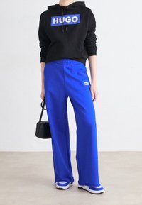 Mustat hupparit, jossa on "HUGO"-logo, siniset collegehousut ja valkoisilla yksityiskohdilla varustetut lenkkarit. Malli pitää kädessään pientä mustaa laukkua. Lattia on vaaleanbeige.