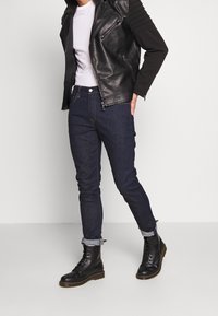 Giacca biker in pelle nera, t-shirt bianca, jeans slim-fit blu scuro, risvolti arrotolati e stivali neri con lacci e suola spessa.