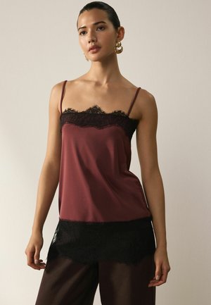 Woman wearing a burgundy camisole with black lace trim and gold hoop earrings, standing against a plain light background.