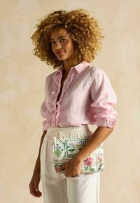 Kreemikujuline lilleline kott, millel on erksad tikitud mustrid, hoitakse käes. Mudel kannab heledat roosa volangiga särki ja valgeid pükse.