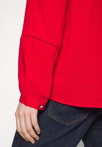 Blouse rouge avec un motif texturé, manches longues et poignet à boutons. Associée à un jeans en denim foncé avec des coutures contrastées.