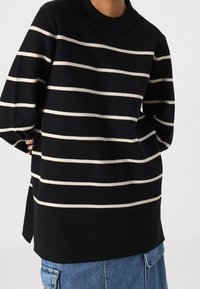 Personne portant un pull noir à rayures blanches horizontales et un pantalon en denim bleu, les mains positionnées derrière le dos.