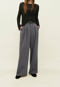 Pull en maille noire transparente associé à un pantalon plissé gris taille haute et une ceinture noire. Tissu lisse, coupe large aux jambes, et chaussures plates noires.