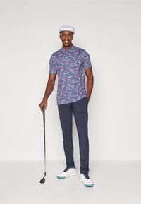 Poloko s krátkymi rukávmi, modré s kvetinovým vzorom a ružovými a bielymi akcentmi, v kombinácii s námorníckymi nohavicami. Biele golfové topánky s námorníckymi šnúrkami, drží golfovú palicu.