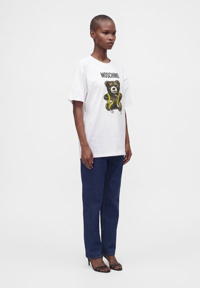 Άτομο που φοράει λευκό t-shirt Moschino με γραφικό αρκουδάκι, σκούρο μπλε τζιν και μαύρα σανδάλια με τακούνι, στέκεται σε απλό λευκό φόντο.