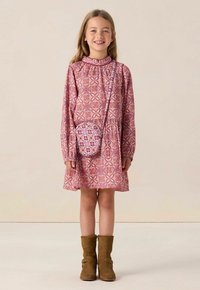 Rosa gemustertes Kleid mit langen Ärmeln, Rüschenausschnitt und einem ausgestellten Rock. Braune Stiefeletten und eine kleine passende Umhängetasche.