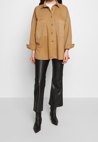 Camicia oversize color tan con due tasche frontali, maniche arrotolate, abbinata a pantaloni in pelle nera e stivali neri a punta.